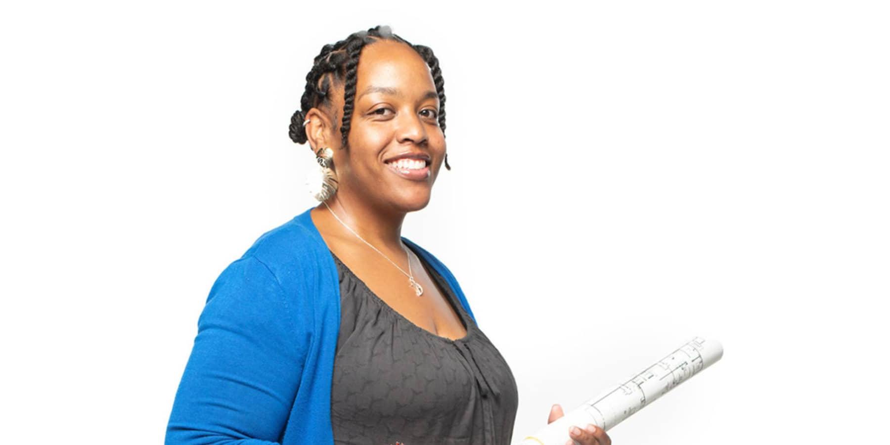 Professor of Interior Architectural Design Azizi Arrington-Slocum, a smiling Black woman with black braided hair, wearing a grey shirt with a blue jacket, and holding a set of rolled-up architectural designs.