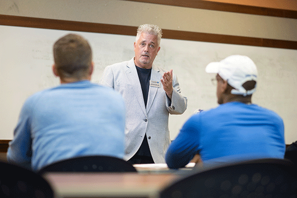 A white male professor stands at the front of a classroom. With white spiked hair, he wears a white dress suit with a dark blue/black undershirt. His right hand is raised toward two male students sitting at desks in front of him. On the left is a white male student wearing a blue long-sleeved shirt and a white baseball cap. On the right is a white male student wearing a blue long-sleeved shirt. A whiteboard is in the background.